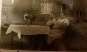 Historisches Foto: zwei Personen sitzen an einem gedeckten Tisch mit Blumenvase vor einem Haus